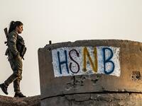 A member of the Bethnahrin Women Protection Forces (HSNB), an all-female Syriac-Assyrian paramilitary group under the umbrella of the Syrian Democratic Forces (SDF), walks past graffiti reading out the group's initials in Latin characters during a commemoration of the fourth anniversary of their creation, in the countryside of the town of Tall Tamr in the northwestern Syrian province of Hasakah, on August 30, 2019.  Delil SOULEIMAN / AFP
