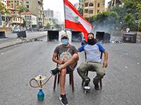 A masked man smokes a waterpipe (also known as shisha or hookah) while another holds up a Lebanese national flag as they sit together before overturned dumpsters blocking a road in the centre of the Lebanese capital Beirut on October 18, 2019 amidst ongoing protests against dire economic conditions. Public anger has simmered since parliament passed an austerity budget in July to help trim a ballooning deficit and flared on October 17 over new plans to tax calls on messaging applications such as WhatsApp, fo