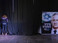 A woman stands in front of curtains at a stage near a sign showing the face of the Blue and White (Kahol Lavan) alliance's leader Benny Gantz, with text in Hebrew reading "we must move forward", at the alliance's electoral headquarters in the coastal city of Tel Aviv early on March 3, 2020. GALI TIBBON / AFP