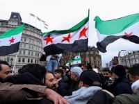 متظاهرون يرفعون أعلام الثورة السورية في ساحة عامة وسط تجمع حاشد، مع مبانٍ تاريخية في الخلفية.