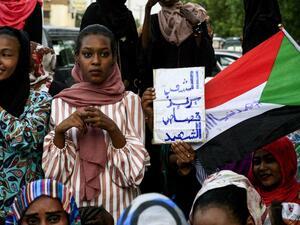 A Sudanese woman holds up a sign reading in Arabic "the people want retribution for the martyrs" next to a waving national flag defaced with the Arabic text "#CiviliaNow". (AFP/ File Photo)