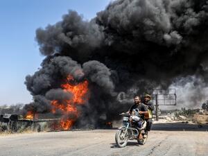 Men ride a motorcycle past a plume of smoke from a blaze at a vehicle gathering point for civilians fleeing from the south of Idlib province, which was hit by reported government forces' bombardment (AFP)