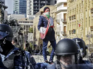 An anti-government protester wearing a Lebanese national flag as a face bandana stands atop a concrete block behind riot police and ahead of a security barrier during a demonstration in the centre of the capital Beirut on January 27, 2020. JOSEPH EID / AFP