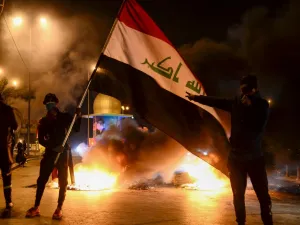Masked anti-government protester wave the national flag before flaming tyres at a make-shift roadblock in the central Iraqi holy shrine city of Najaf on February 1, 2020. Iraq's president named former communications minister Mohammad Allawi as the country's new prime minister after an 11th-hour consensus among political blocs, but the streets seemed divided on his nomination. Haidar HAMDANI / AFP