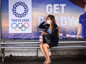 A woman wearing a facemask, amid concerns over the spread of the COVID-19 coronavirus, sits at a bus stop in front of a Tokyo 2020 Olympics advertisement in Bangkok on March 16, 2020. Mladen ANTONOV / AFP