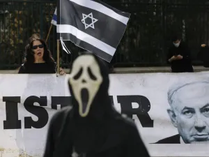 A person wearing a ghost-face mask walks past an Israel woman holding her national flag in black colour motifs, standing behind a portrait of Prime Minister Benjamin Netahyahu, during a protest outside the Knesset (parliament) in Jerusalem on March 23, 2020 against the recent decision by Knesset speaker to shut it down, and against what they describe as "damaged to Israel's democracy". MENAHEM KAHANA / AFP