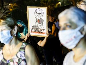 A protester, mask-clad due to the COVID-19 coronavirus pandemic, stands holding a sign showing a cartoon depicting Israeli Prime Minister Benjamin Netanyahu stepping on text dripping with red drops reading in Hebrew "democracy", during a demonstration against the Israeli government in the Mediterranean coastal city of Tel Aviv on August 1, 2020. JACK GUEZ / AFP
