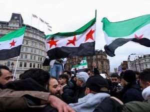 متظاهرون يرفعون أعلام الثورة السورية في ساحة عامة وسط تجمع حاشد، مع مبانٍ تاريخية في الخلفية.