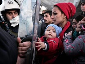 A woman holds a baby as migrants and refugees stand facing Greek police while waiting to cross into Macedonia on March 2, 2016, where thousands of people are stranded. (AFP/Louisa Gouliamaki)