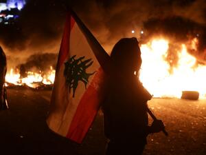 An Lebanese anti-government protester carries a Lebanese flag in front of burning tires blocking the main highway linking the city of Tripoli to Beirut at the coastal city of Byblos, north of the capital on November 13, 2019.  (AFP/ File Photo)