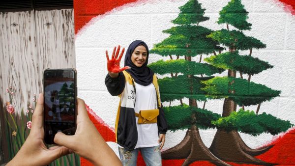 A woman shows her red-painted hand as she poses for a picture next to a mural depicting two cedars during a demonstration in southern city of Sidon on the seventh day of protest against tax increases and official corruption, on October 23, 2019. Mahmoud ZAYYAT / AFP