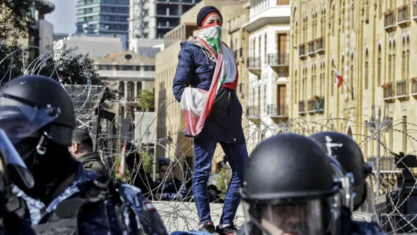 An anti-government protester wearing a Lebanese national flag as a face bandana stands atop a concrete block behind riot police and ahead of a security barrier during a demonstration in the centre of the capital Beirut on January 27, 2020. JOSEPH EID / AFP