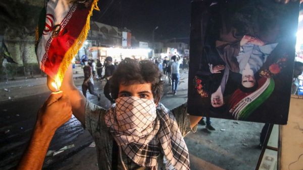 In this file photo taken on September 07, 2018, an Iraqi protester holds up an Iranian flag as another sets it on fire for him, next to a portrait depicting Iran's former and current Supreme Leaders Ayatollah Khomeini and Khamenei, during demonstrations against the government and the lack of basic services in Basra. Mocking clerics, falling in love at rallies and mending a broken society: even if Iraq's young protesters have failed to overthrow entrenched politicians, they have scored by shattering decades-