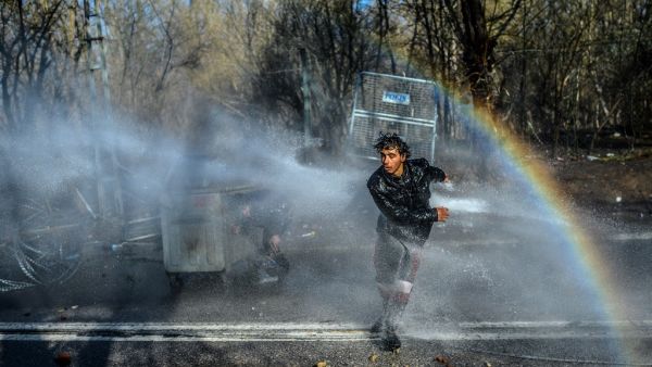 A migrant runs away as another one takes cover behind a trash container as Greek police uses water cannons to block them trying to break fences in the Turkey-Greece border province of Edirne on March 7, 2020 as makeshift camps have sprung up around the border where thousands of refugees have been encouraged by Turkey to leave for the European Union, in a bid to gain Western backing in Syria. As part of the 2016 agreement, Turkey agreed to stop the flow of migrants to Europe in exchange for billions of euros