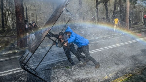 Migrants take cover behind a wooden board as Greek police uses water cannons to block them trying to break fences in the Turkey-Greece border province of Edirne on March 7, 2020 as makeshift camps have sprung up around the border where thousands of refugees have been encouraged by Turkey to leave for the European Union, in a bid to gain Western backing in Syria. As part of the 2016 agreement, Turkey agreed to stop the flow of migrants to Europe in exchange for billions of euros, but Ankara accused the Europ