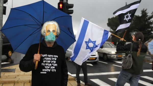 A man holding an umbrella, wearing a protective face mask beneath another mask showing the face of late physicist Albert Einstein, and a hoodie sweater quoting a verse from the biblical book of Isaiah, while another waves an Israeli flag and another Israeli flag with the colours inverted during a protest outside the Knesset (parliament) in Jerusalem on March 19, 2020. AFP/File