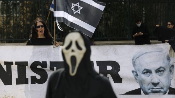 A person wearing a ghost-face mask walks past an Israel woman holding her national flag in black colour motifs, standing behind a portrait of Prime Minister Benjamin Netahyahu, during a protest outside the Knesset (parliament) in Jerusalem on March 23, 2020 against the recent decision by Knesset speaker to shut it down, and against what they describe as "damaged to Israel's democracy". MENAHEM KAHANA / AFP