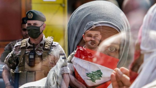 This double exposure photo taken on May 3, 2020 shows a Lebanese protester wearing a Lebanese flag face mask as members of the Lebanese Armed Forces stand guard during a demonstration against dwindling economic conditions, at al-Nour Square in the centre of the northern port city of Tripoli. Ibrahim CHALHOUB / AFP