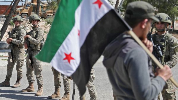 A demonstrator holds a flag of the Syrian opposition as he walks before Turkish soldiers securing a section of the M4 highway, which links the northern Syrian provinces of Aleppo and Latakia, near Ariha in Syria's jihadist-controlled northwestern Idlib province on May 12, 2020. OMAR HAJ KADOUR / AFP