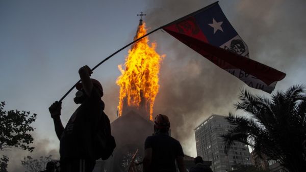 A demonstrator flutters a Chilean flag outside the burning church of Asuncion, set on fire by protesters, on the commemoration of the first anniversary of the social uprising in Chile, in Santiago, on October 18, 2020, as the country prepares for a landmark referendum. MARTIN BERNETTI / AFP
