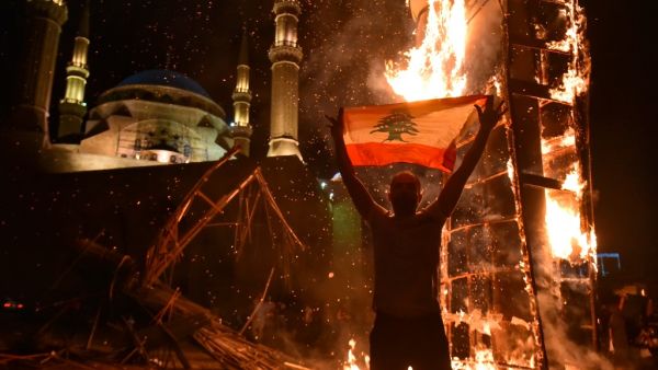 A Lebanese man raises a national flag as the ‘Revolution fist’, symbol of Lebanon’s October 2019 uprising, burns after being torched during clashes between anti-government protesters and supporters of former prime minister Saad Hariri, in the capital Beirut's central Martyr's square, on October 21, 2020. Hariri resigned as premier in October 2019 in the wake of unprecedented street protests, but he is now expected to make a comeback at the helm of the next government. Most parliamentary blocs have pledged s
