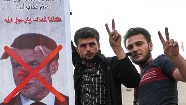 Men flash the victory gesture as they pose for a picture next to a sign condemning French President Emmanuel Macron, and depicting him with a pig snout, for his comments over Prophet Mohammed cartoons in Syria's rebel-held northwestern city of Idlib on October 25, 2020. OMAR HAJ KADOUR / AFP