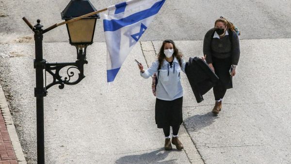 Israeli schoolgirls, clad in mask due to the COVID-19 coronavirus pandemic, walk underneath an Israeli flag hanging from a lamp post along al-Shuhada street, largely closed to Palestinians, in the city centre of the flashpoint city of Hebron in the occupied West Bank on February 21, 2020. HAZEM BADER / AFP