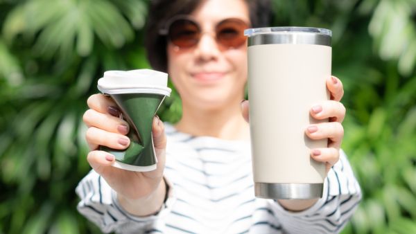 Woman holds takeaway coffee cup in both hands, one is a single use paper cup with plastic lid, the other one is a reusable stainless tumbler