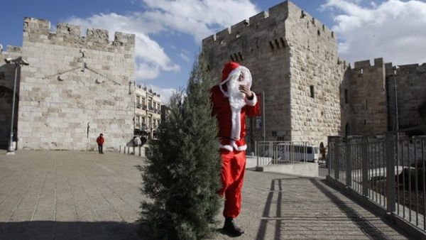 Issa Kassissieh, dressed as Santa Claus, carries a Christmas tree outside the Jaffa Gate in Jerusalem. [mondoweiss]