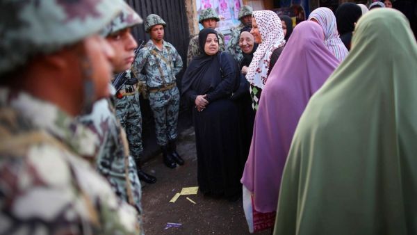 Women standing near The Citadel wait in line to vote watched by soldiers.