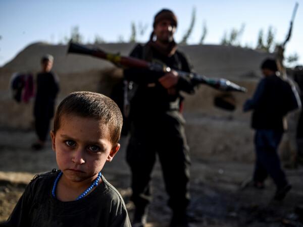 In this photo taken on September 27, 2020, a young boy looks at the camera as a policeman holding a rocket-propelled grenade (RPG) stands behind in a house at Deh Qubad village in Maiwand district of Kandahar province. The dry and dusty village of Aziz Abad in Maiwand district sits on the frontline of Afghanistan's war and is currently under government control -- but only just. WAKIL KOHSAR / AFP