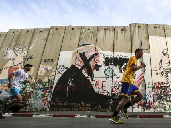 Participants run past a mural depicting U.S. President Donald Trump drawn along Israel's controversial separation barrier, which divides the West Bank from Jerusalem, in the biblical town of Bethlehem during the 6th International Palestine Marathon on Mar. 23, 2018. 
(Musa AL SHAER / AFP)