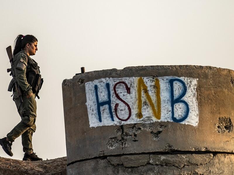 A member of the Bethnahrin Women Protection Forces (HSNB), an all-female Syriac-Assyrian paramilitary group under the umbrella of the Syrian Democratic Forces (SDF), walks past graffiti reading out the group's initials in Latin characters during a commemoration of the fourth anniversary of their creation, in the countryside of the town of Tall Tamr in the northwestern Syrian province of Hasakah, on August 30, 2019.  Delil SOULEIMAN / AFP