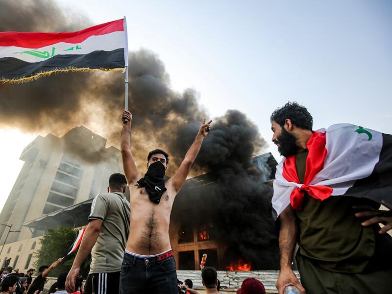 A topless masked protester flashes the victory gesture as he waves an Iraqi national flag before a burning building during a demonstration against state corruption and poor services, between the capital Baghdad's Tahrir Square and the high-security Green Zone district, on October 1, 2019.  AHMAD AL-RUBAYE / AFP