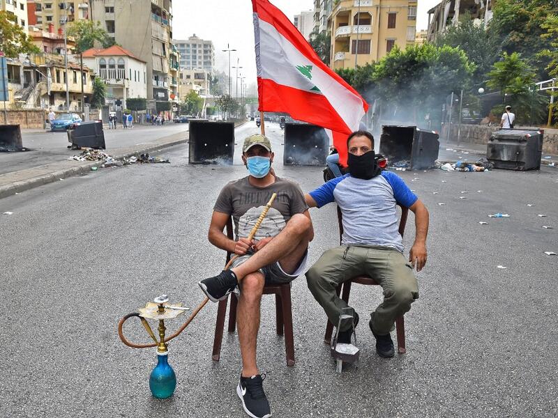 A masked man smokes a waterpipe (also known as shisha or hookah) while another holds up a Lebanese national flag as they sit together before overturned dumpsters blocking a road in the centre of the Lebanese capital Beirut on October 18, 2019 amidst ongoing protests against dire economic conditions. Public anger has simmered since parliament passed an austerity budget in July to help trim a ballooning deficit and flared on October 17 over new plans to tax calls on messaging applications such as WhatsApp, fo