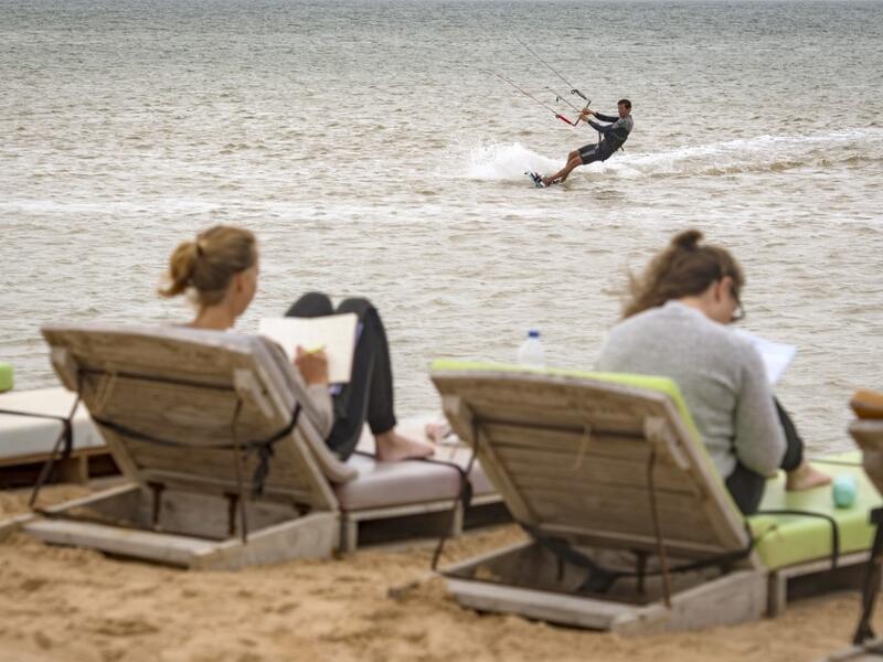 A picture taken on October 10, 2019, shows tourists reading as a kitesurfer glides on waves at Dakhla beach in Morocco-administered Western Sahara. In the heart of disputed Western Sahara, a former garrison town has become an unlikely tourist magnet after kitesurfers discovered the windswept desert coast on the Atlantic is perfect for their sport. FADEL SENNA / AFP