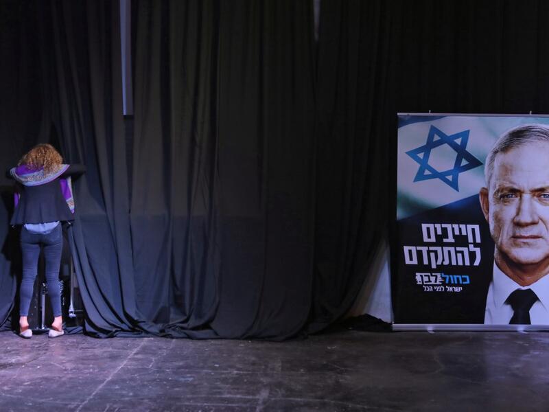 A woman stands in front of curtains at a stage near a sign showing the face of the Blue and White (Kahol Lavan) alliance's leader Benny Gantz, with text in Hebrew reading "we must move forward", at the alliance's electoral headquarters in the coastal city of Tel Aviv early on March 3, 2020. GALI TIBBON / AFP