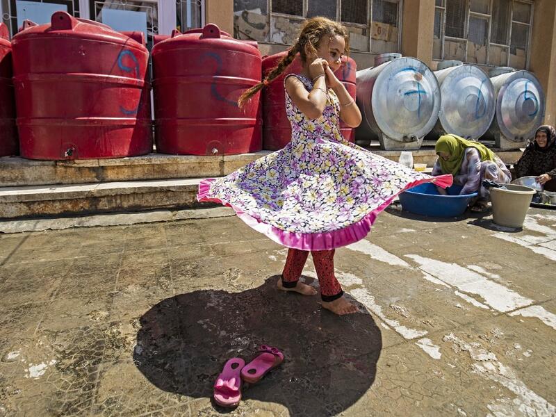 A girl walks past women washing pots and pans by cisterns at the yard of a school building where Syrians -- displaced from the area of Ras al-Ain by the Turkish offensive on the northeast -- are staying in the city of Hasakah, on June 30, 2020. Delil SOULEIMAN / AFP