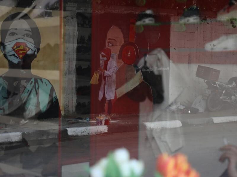 The image of a Palestinian student painting a large mural of a mask-clad girl is reflected on the doors of a shop in Gaza City on October 22, 2020, amid the novel coronavirus pandemic crises. (Instagram)