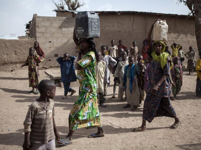 Two women carrying jerrycans filled with water walk past children in the town of Banki in northeastern Nigeria on April 26, 2017. (AFP/Florian Plaucheur)