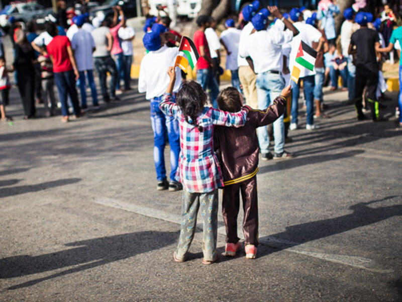 A brother and sister wave flags and watch as planes from the Royal Jordanian Air Force fly overhead in the air show.