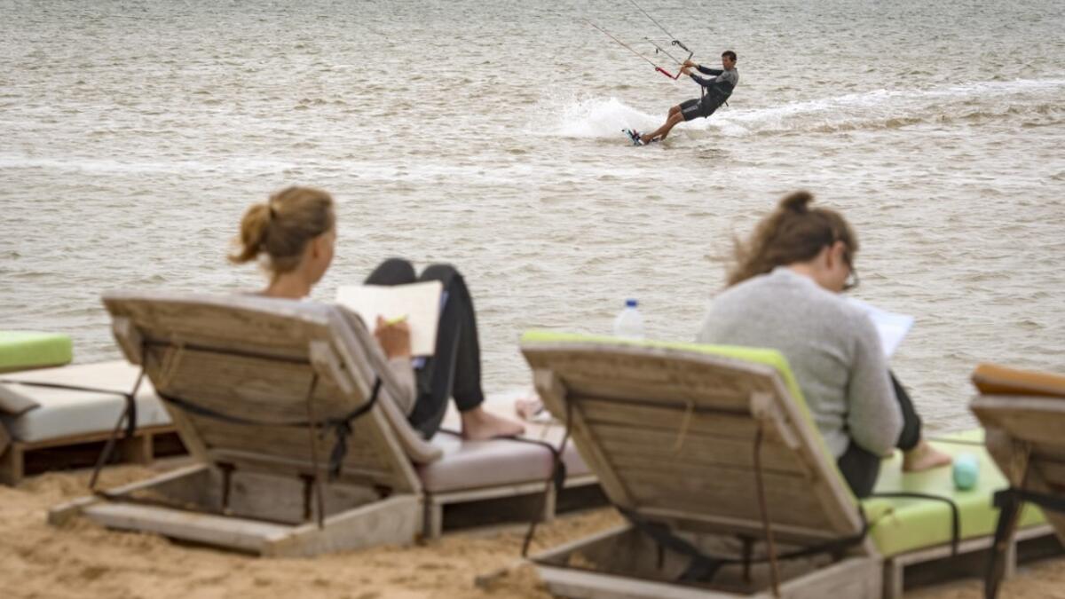 A picture taken on October 10, 2019, shows tourists reading as a kitesurfer glides on waves at Dakhla beach in Morocco-administered Western Sahara. In the heart of disputed Western Sahara, a former garrison town has become an unlikely tourist magnet after kitesurfers discovered the windswept desert coast on the Atlantic is perfect for their sport. FADEL SENNA / AFP