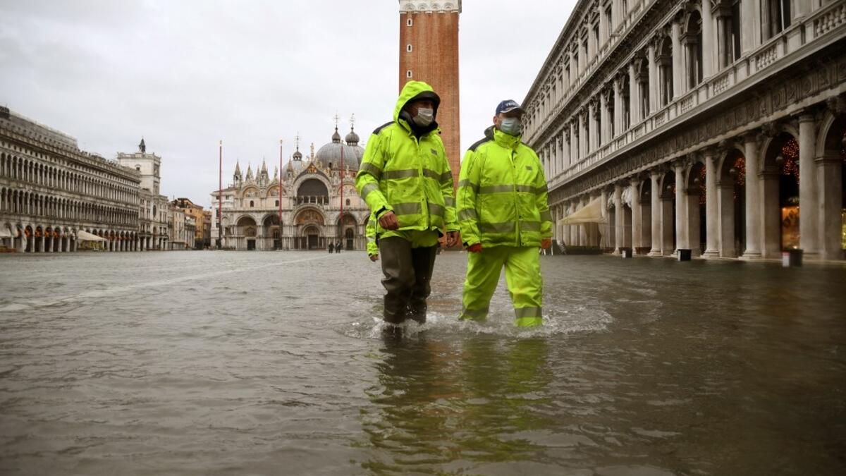 Men wearing waterproof gear walk across a flooded St. Mark's Square on December 8, 2020 following a high tide "Alta Acqua" event following heavy rains and strong winds, and the mobile gates of the MOSE Experimental Electromechanical Module that protects the city of Venice from floods, were not lifted. ANDREA PATTARO / AFP