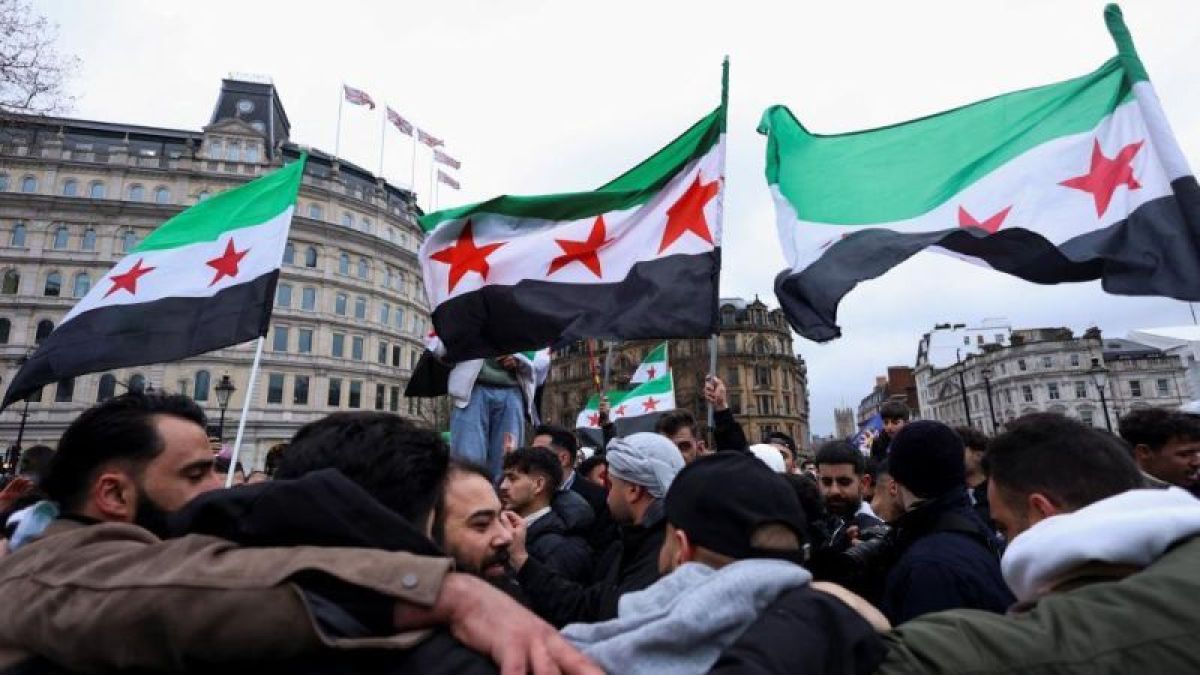 متظاهرون يرفعون أعلام الثورة السورية في ساحة عامة وسط تجمع حاشد، مع مبانٍ تاريخية في الخلفية.