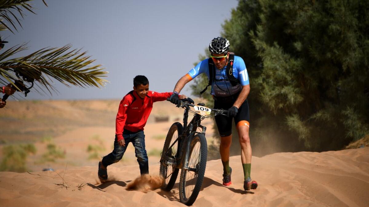 A boy pushes a participant's bike over a sand dune during the Stage 1 of the 14th edition of Titan Desert 2019 mountain biking race around Merzouga in Morocco on April 28, 2019.  FRANCK FIFE / AFP